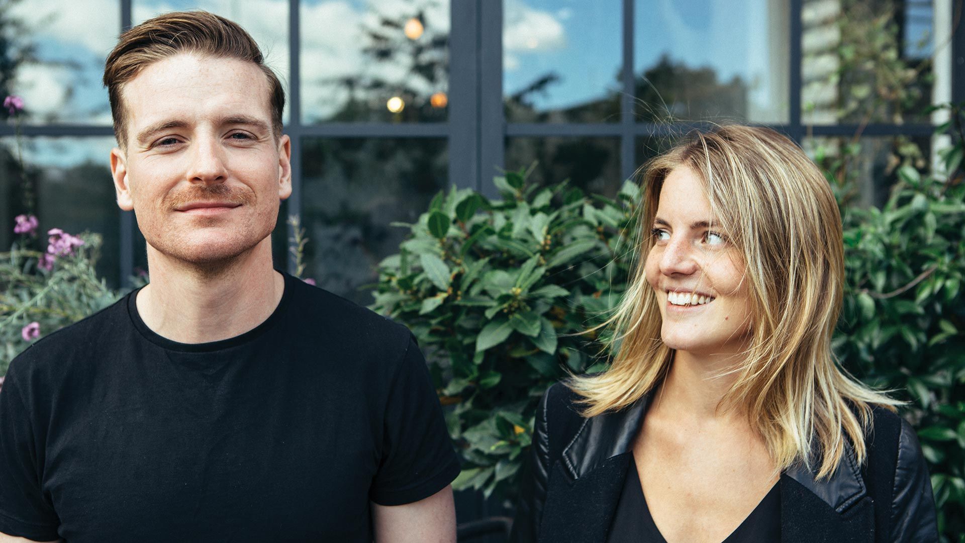 Photo of a woman smiling knowingly to a man, who in turns is smiling to camera.